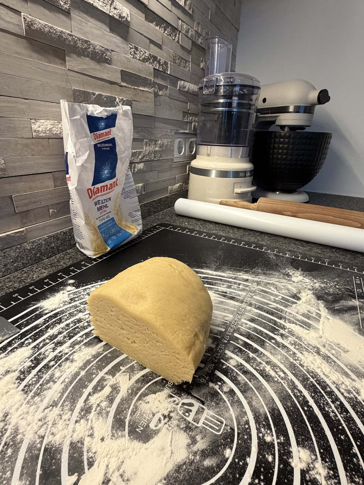 Butterplätzchenteig auf einer bemehlten Backmatte neben Teigrolle und Küchenmaschine